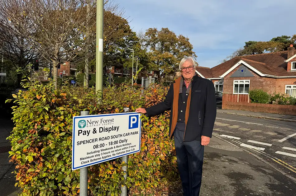 An older man with white hair and glasses stands next to a Pay & Display car park sign on Spencer Road South, with houses, bushes, and a sunny blue sky in the background.