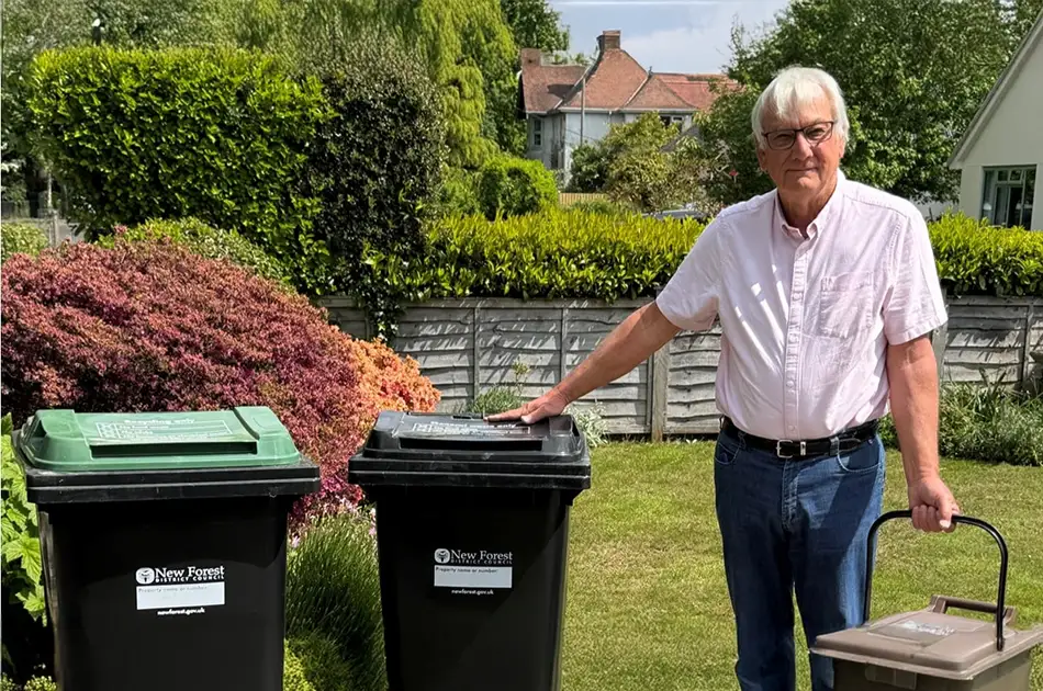 An older man in a pink shirt and blue jeans stands in a garden, holding a small brown food waste bin and resting his hand on one of two large black recycling bins with green lids. a wooden fence and greenery are in the background.