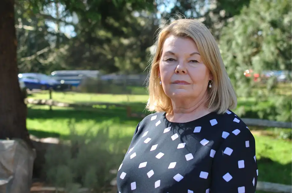 A middle aged woman with blonde hair wearing a black top with white square patterns stands outdoors in a sunny park with green grass and trees in the background.