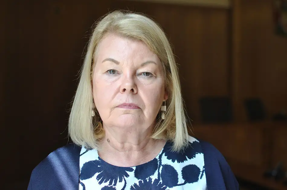 A woman with straight blonde hair and light skin wears a navy and white patterned top, looking directly at the camera with a neutral expression. the background is softly blurred and appears to be indoors with wooden walls.