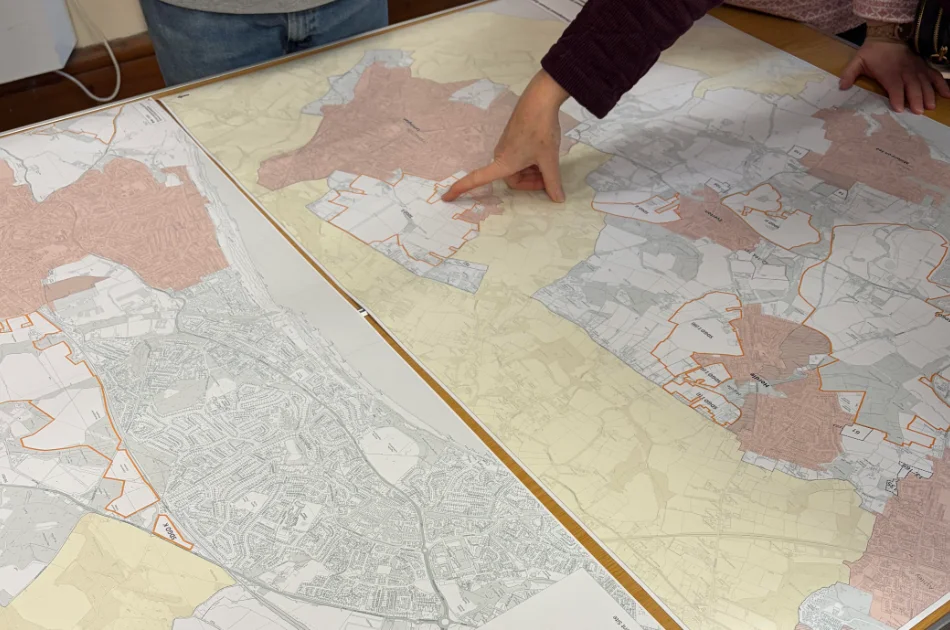 Two people stand over large, detailed maps spread on a table, with one person pointing to a specific area outlined in orange on the central map. The maps show different regions and zoning areas.