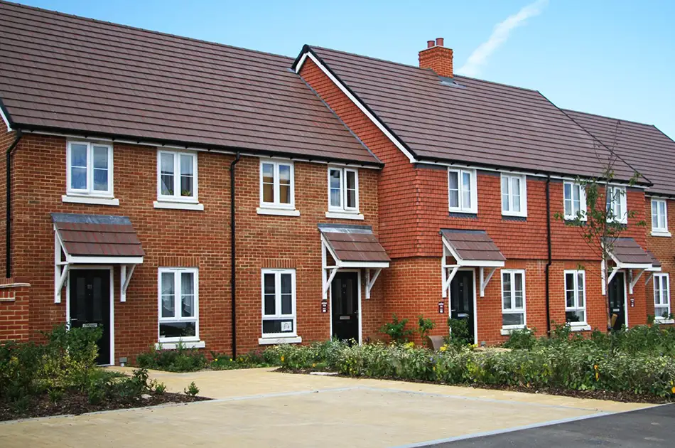 A row of newly built red brick terraced houses with brown tiled roofs, small front gardens, and black front doors, set against a clear blue sky.