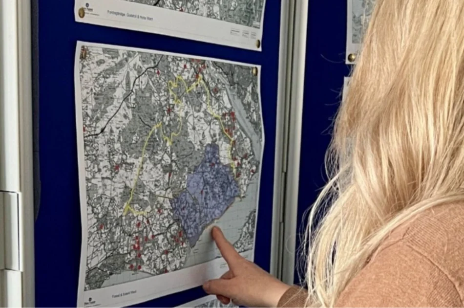 A person with long blonde hair points at a section of a detailed map attached to a blue display board, examining the area closely.