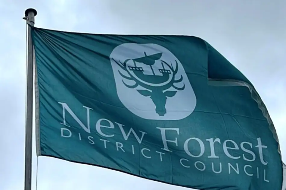 A flag for new forest district council flutters in the wind, featuring a white emblem of a stag’s head with a bird on its antlers above the council’s name on a teal background.