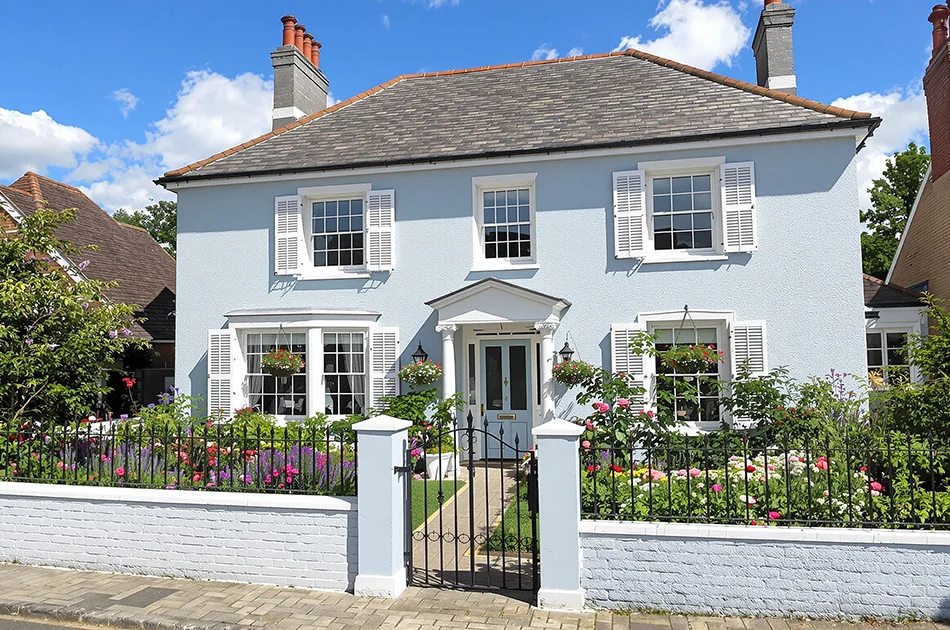 A charming two-story light blue house with white shutters, a lush flower garden, black iron gate, and a sunny, clear sky backdrop.