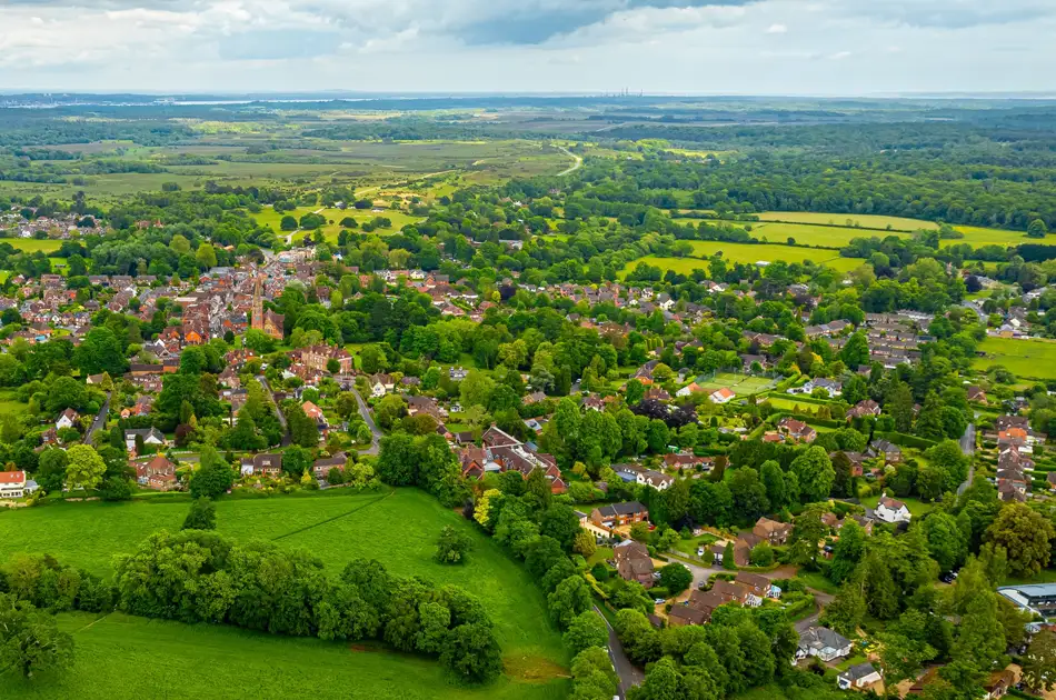 Aerial view of a small village surrounded by lush green fields, trees, and countryside under a partly cloudy sky. Rows of houses and narrow roads are visible among the greenery.