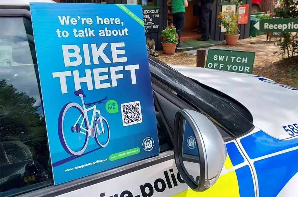 A blue sign on a police car reads, we're here to talk about bike theft with an image of a bicycle, a qr code, and police logos. a green reception sign and potted plants are visible in the background.
