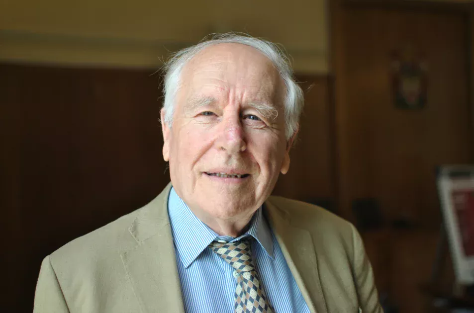 An older man with white hair, wearing a beige jacket, blue striped shirt, and patterned tie, smiles gently at the camera. The background is softly lit with wooden walls and a blurred framed picture.